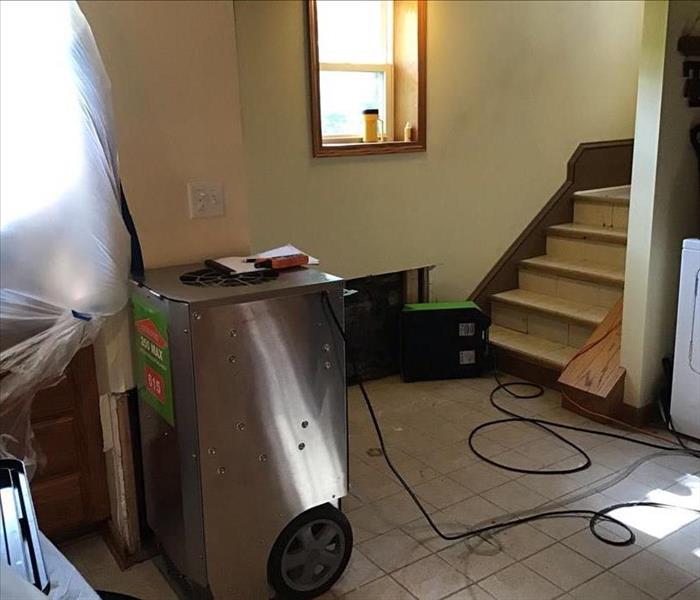 Silver and green dehumidifier on a tile floor with a stairway and a green air mover on the floor. 