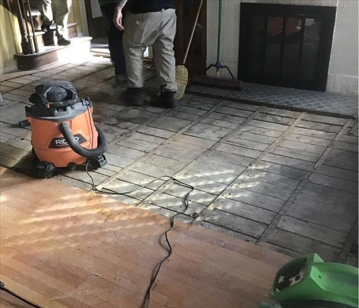 Hard wood floor partially removed in front of a fireplace, with a green air mover and an orange shop vacuum.