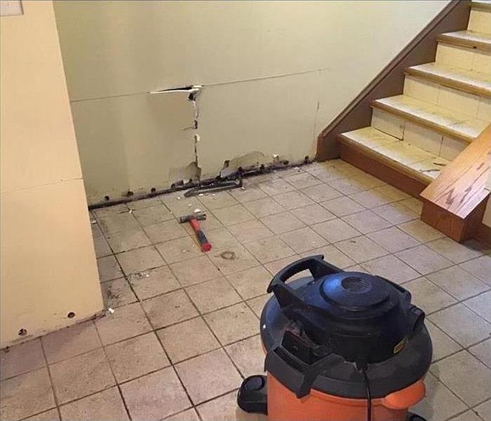 Dirty tile floor with white drywall with holes at the bottom with a stairway and an orange shop vacuum on the floor. 